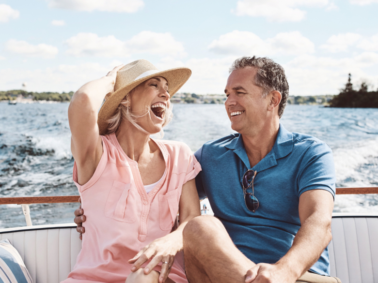 A cheerful couple enjoys a sunny day on a boat. The woman in a pink shirt and straw hat laughs, while the man in a blue shirt smiles back at her.