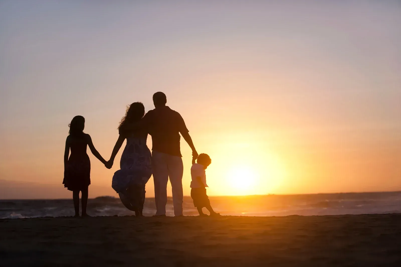 The silhouette of a family of four stands on a sandy beach, holding hands as they face a vibrant sunset over the ocean. The sky displays hues of orange, pink, and gray.