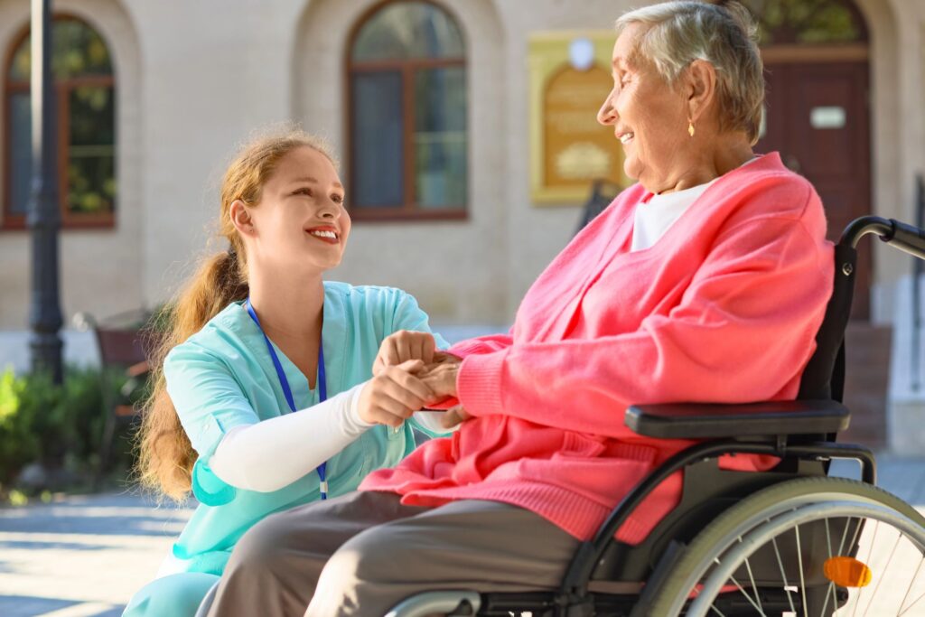 A young caregiver with light brown hair in a ponytail, wearing a teal scrub top and a blue lanyard, is holding the hands of an elderly woman sitting in a wheelchair. The elderly woman has short grey hair, is wearing a white shirt under a coral pink cardigan, and is also smiling. They are outdoors, with a building with arched windows and greenery in the background. The image conveys a sense of care and connection.