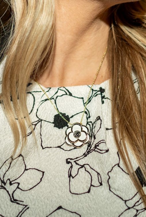 A white flower pendant with a gold-toned outline and interlocking logo hangs from a delicate chain over a floral top. Discover distinctive necklaces at Eliana Curated in Old Town Alexandria, Virginia.