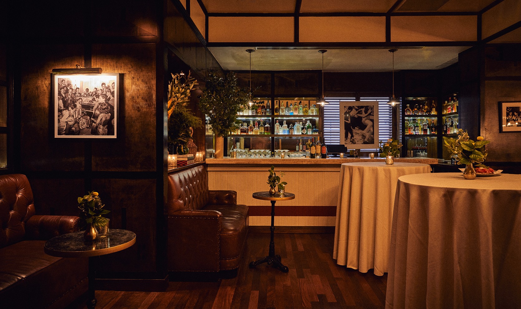 A sophisticated restaurant interior featuring dark wood paneled walls a tufted leather banquette and cocktail tables. The back bar is glowing with bottles creating a warm moody atmosphere.