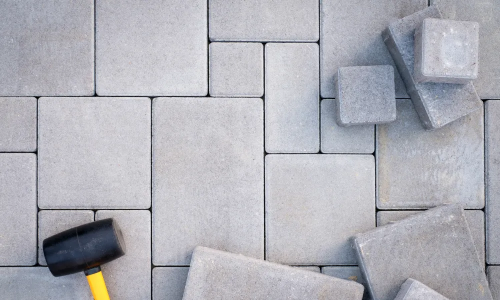 An overhead shot of a collection of gray concrete paving stones laid out in a pattern, with a rubber mallet on the left side of the frame. Several paving stones are stacked loosely in the upper right corner, suggesting a construction or landscaping project.