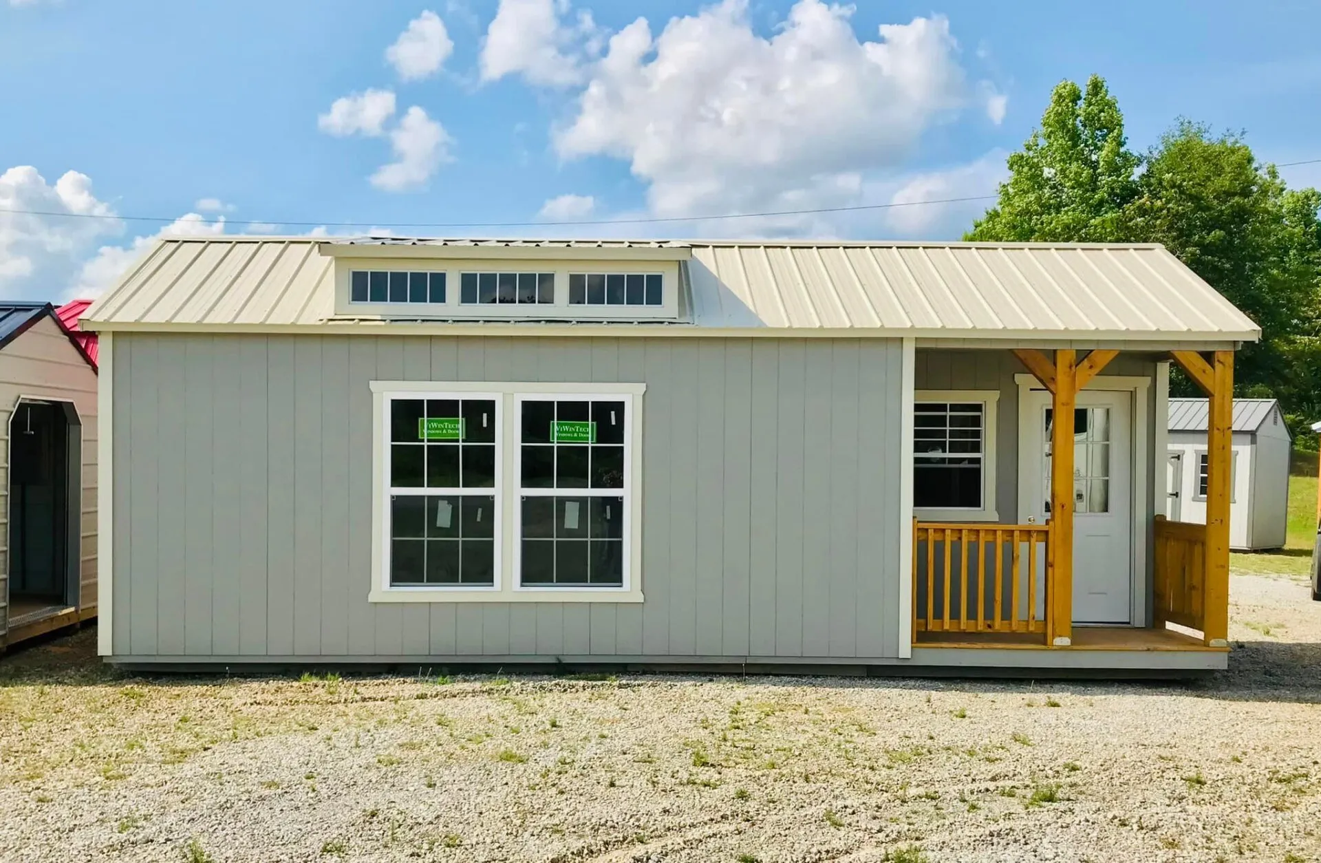 Gray portable cabin with covered front porch, clerestory windows, white trim, and durable metal roof. Designed for comfortable living, office space, or recreational property use with inviting appearance.