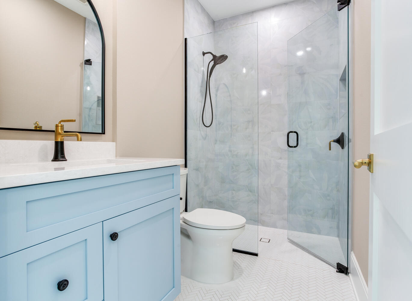 Modern bathroom with a light blue vanity, arched mirror, walk-in glass shower, marble-look tile, and polished gold fixtures.