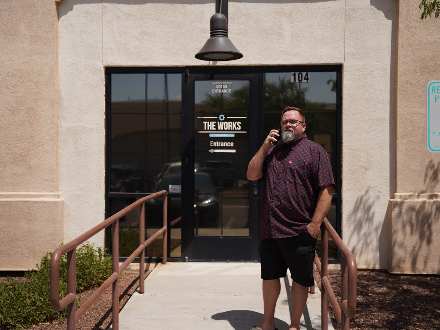 In this image, Mike Rux, a seasoned professional at Ciphers Digital Marketing, is seen outside the Gilbert, Arizona office. He is dressed casually in a patterned short-sleeve shirt and black shorts, standing on a ramp leading to the building entrance marked "The Works." Mike is engaged in a phone conversation, exuding a focused and approachable demeanor. The building's exterior is light-colored, with large glass doors and a prominent lamp above the entrance, capturing the sunny and professional atmosphere of the location.