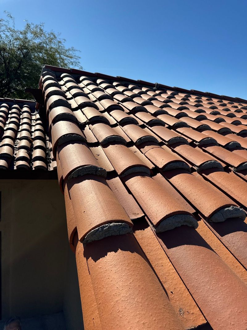 A close-up, low-angle shot highlights a freshly installed red clay barrel tile roof. The image captures the rhythmic texture of the tiles and mortar under a clear, bright blue sky.