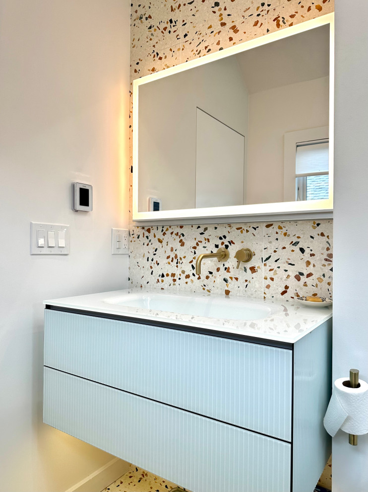 A modern bathroom vanity with a light blue fluted cabinet, a white countertop, and a terrazzo tile backsplash. A large, backlit mirror hangs above the sink, which has gold-toned faucets. The wall features light switches and a thermostat. A roll of toilet paper is mounted on the right side of the vanity.