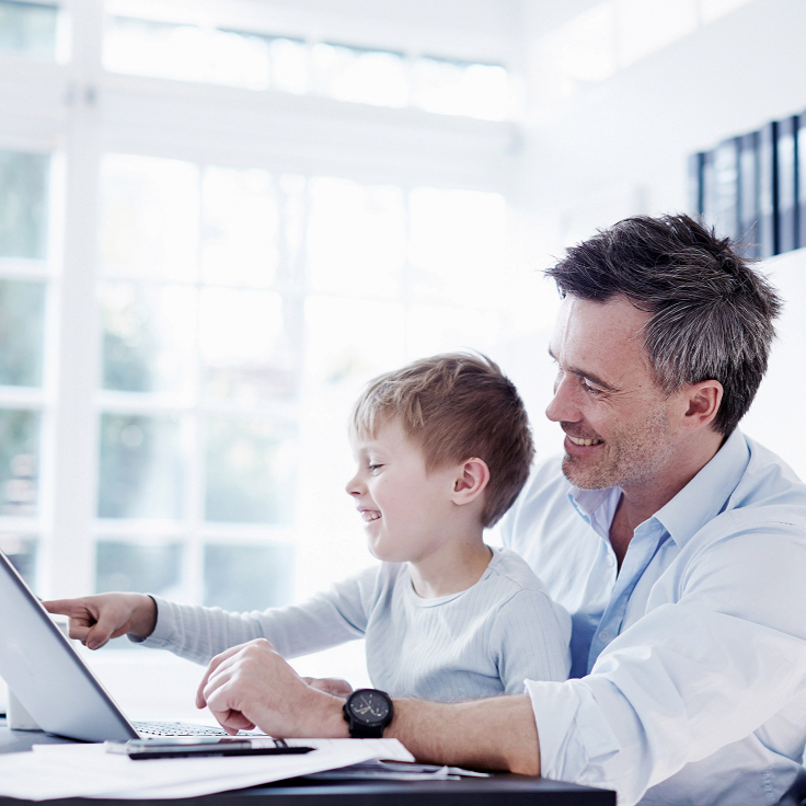 Father and son smiling at a laptop in a bright room with large windows and natural light. The scene conveys happiness and bonding.