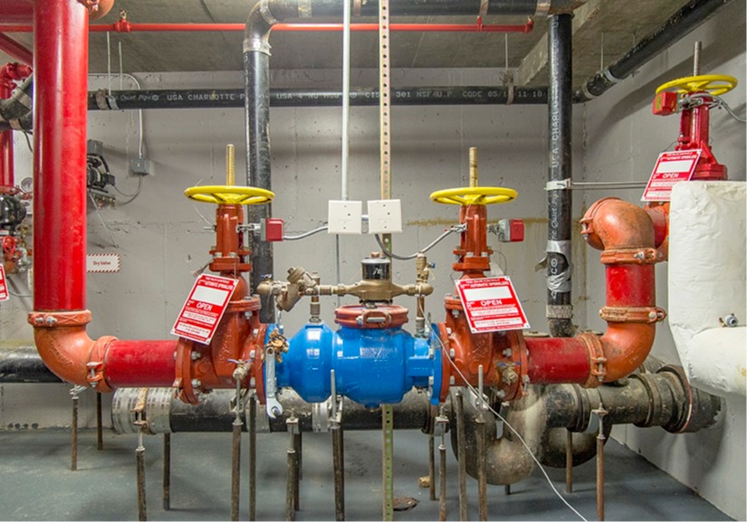 A mechanical room features red and black pipes with yellow valves and red warning tags. Center is a blue valve assembly. The setting appears industrial.