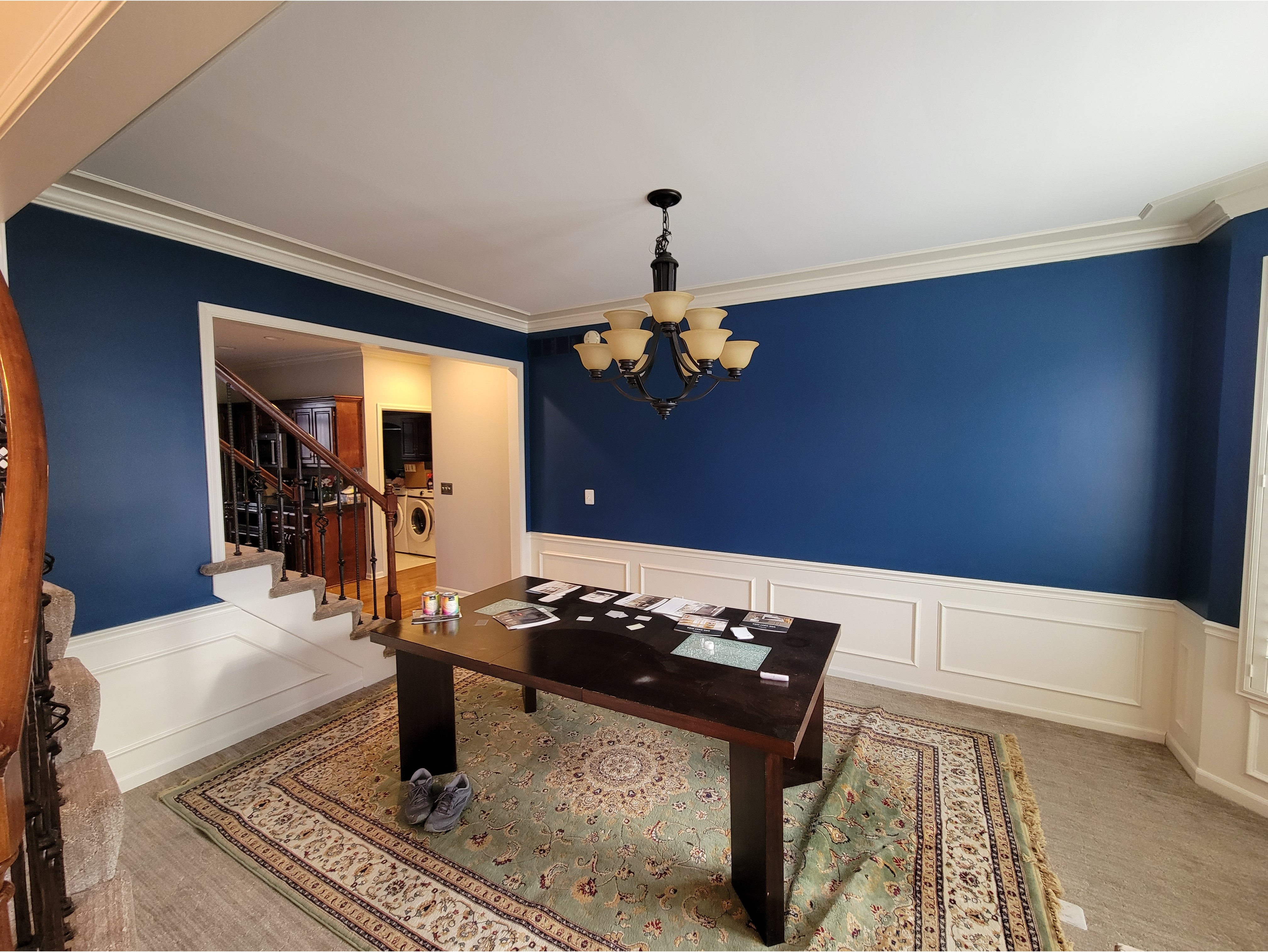 A modern dining room showcasing a fresh interior paint job with a bold navy blue accent wall and white wainscoting for a classic, elegant look. The room features decorative trim molding, a chandelier, and a dark wood table set on a patterned area rug. Perfect for highlighting interior painting services, trim work, and accent wall designs.