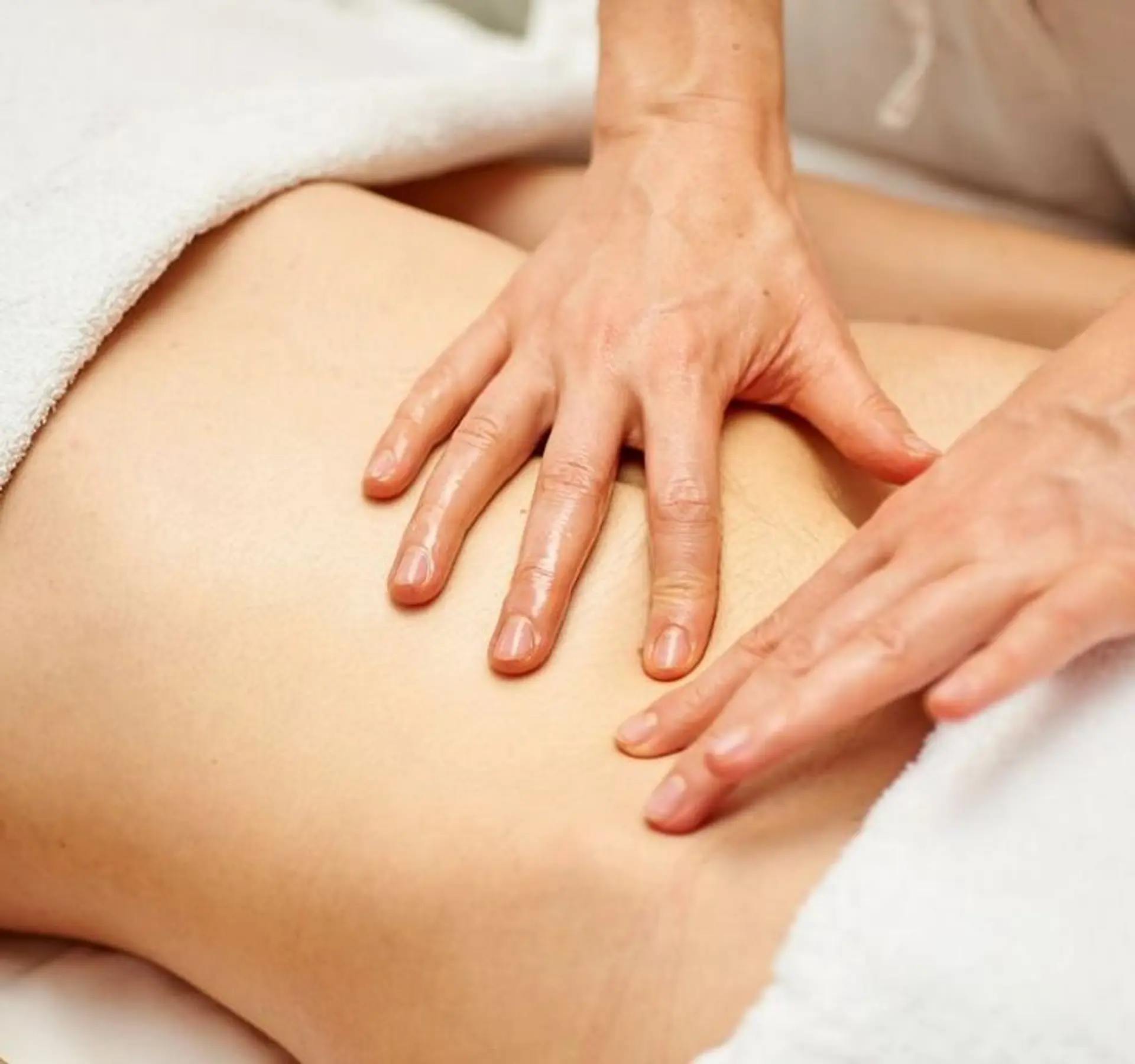 A close-up shot shows two hands massaging the abdomen of a person. The hands are oiled and appear to be performing a therapeutic massage. A white towel is visible on the left side of the frame, suggesting a spa or massage therapy setting.