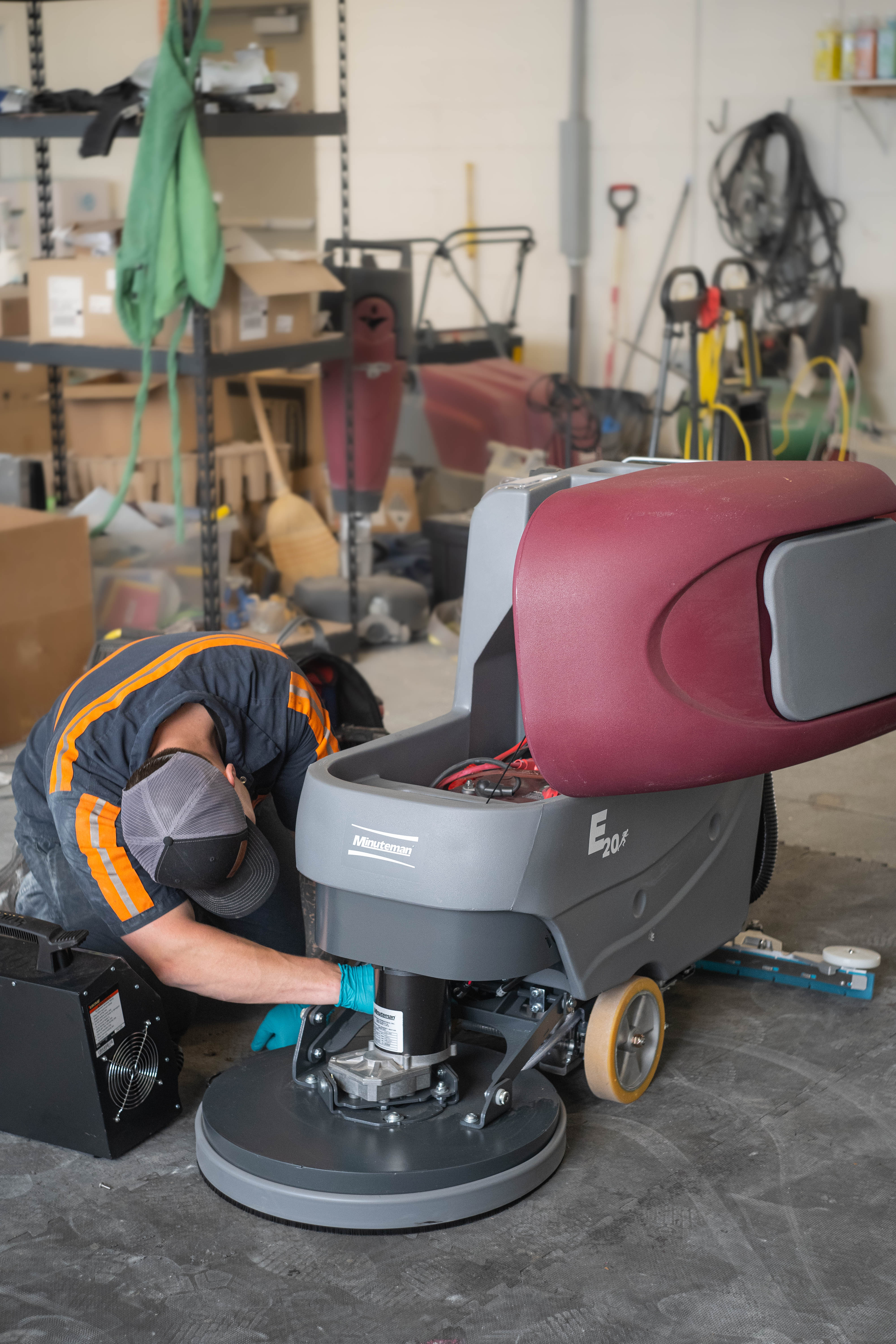 A professional WaySolutions field technician kneels to perform deep mechanical maintenance on the lower scrub deck and motor assembly of a Minuteman E20 walk-behind floor scrubber. Our cross-trained experts in Layton deliver comprehensive ground-level repairs, ensuring heavy-duty commercial cleaning equipment maintains peak scrubbing performance.