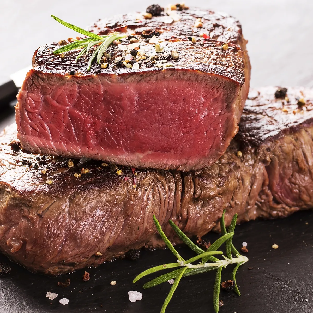A close-up, mouthwatering shot of two perfectly cooked steaks, one sliced to reveal a juicy, medium-rare interior. The steaks are seasoned with coarse salt, black peppercorns, and garnished with fresh rosemary sprigs, presented on a dark slate surface.