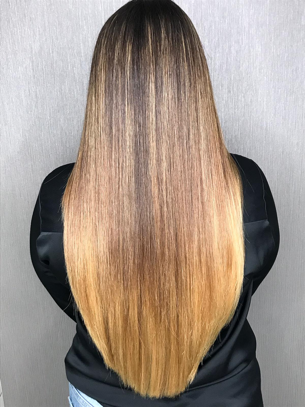 A back view of a person with long, straight, and smoothly styled hair that transitions from dark brown at the roots to a warm blonde at the ends. The person is wearing a black satin-like top and parts of light blue denim jeans are visible at the bottom. The background is a textured gray wall.