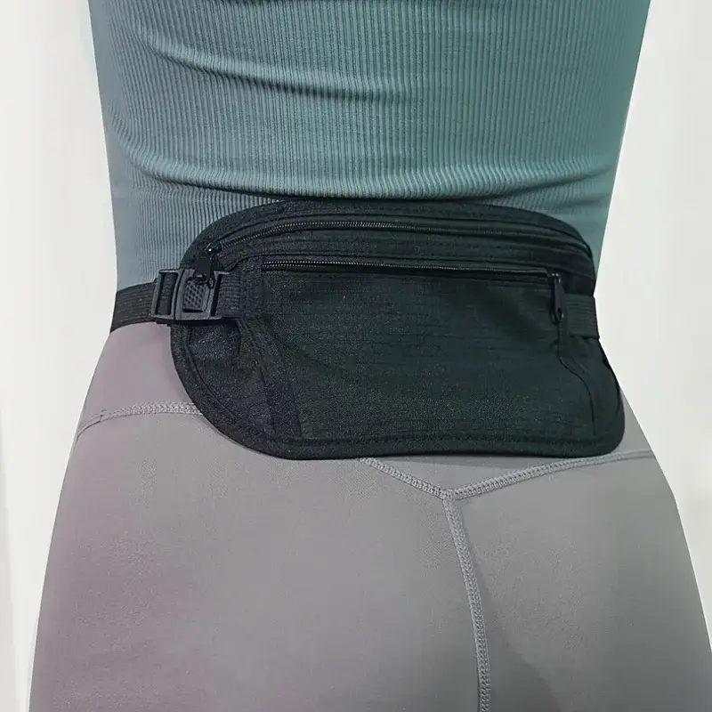 A close-up shot focuses on a person wearing a black fanny pack around their waist. The fanny pack is made of a textured black fabric with two zippers. It is worn over grey leggings and a teal, ribbed tank top. The background is a plain white wall.