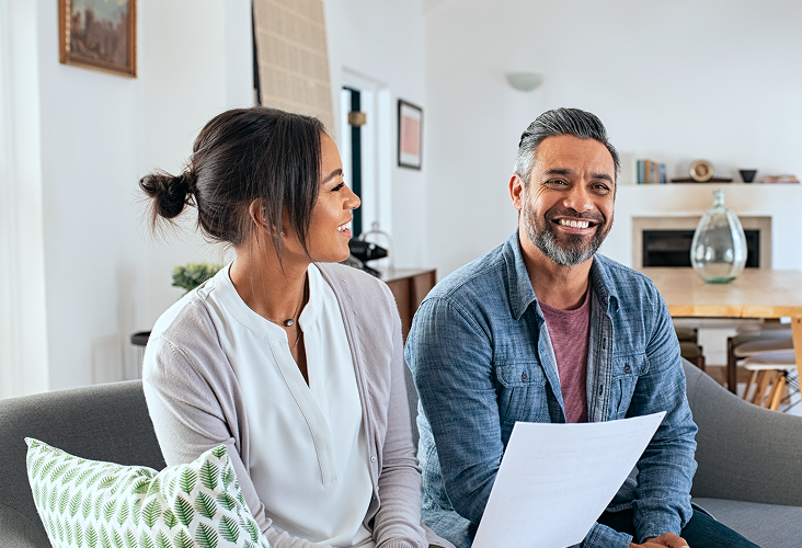 A smiling man and woman sitting in their living room reviewing a document.