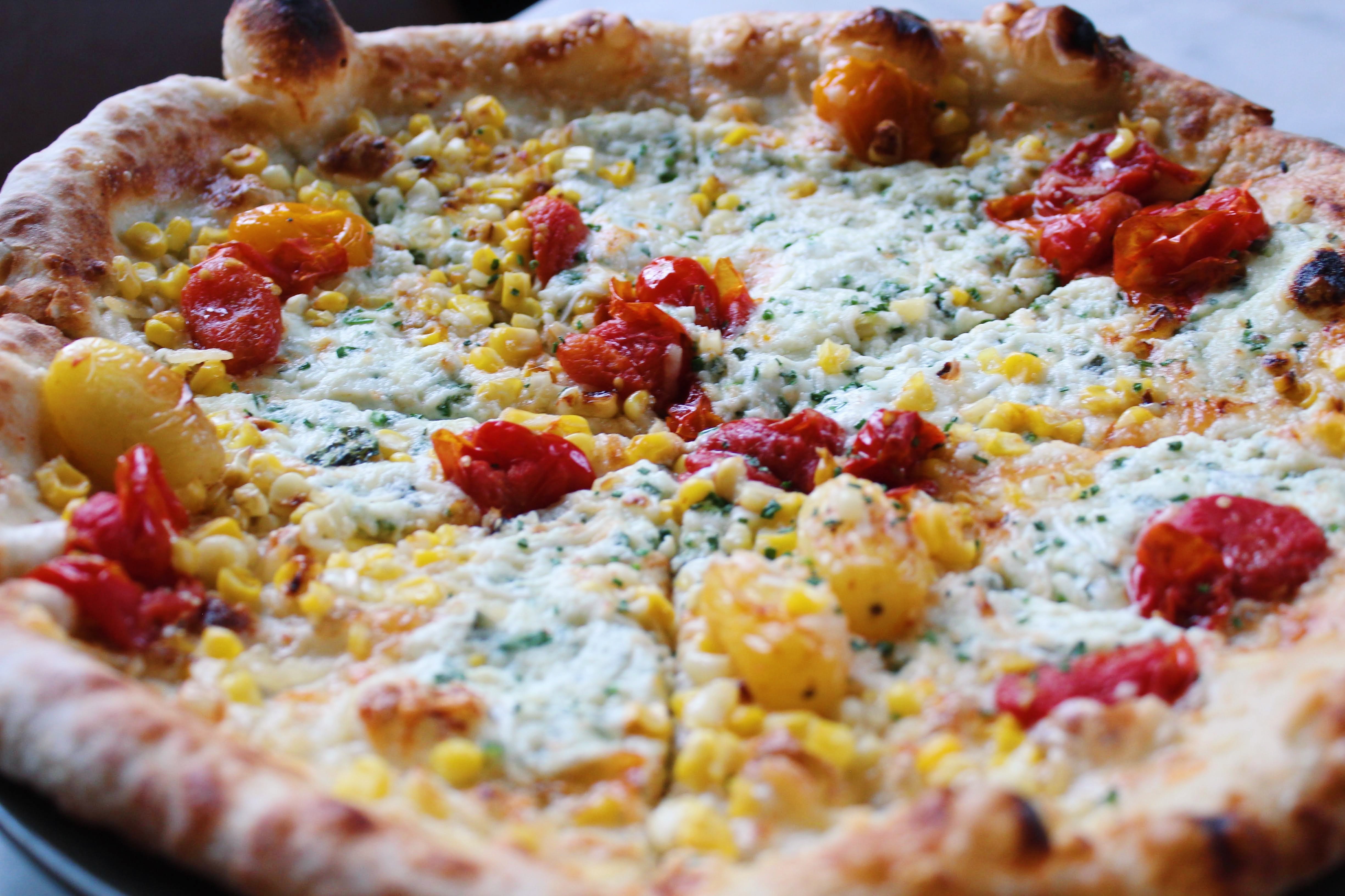 The Summer Bianca — a wood-fired white pizza layered with whipped ricotta, parmesan, and pecorino romano, topped with burst cherry tomatoes, roasted corn, and fresh chives.