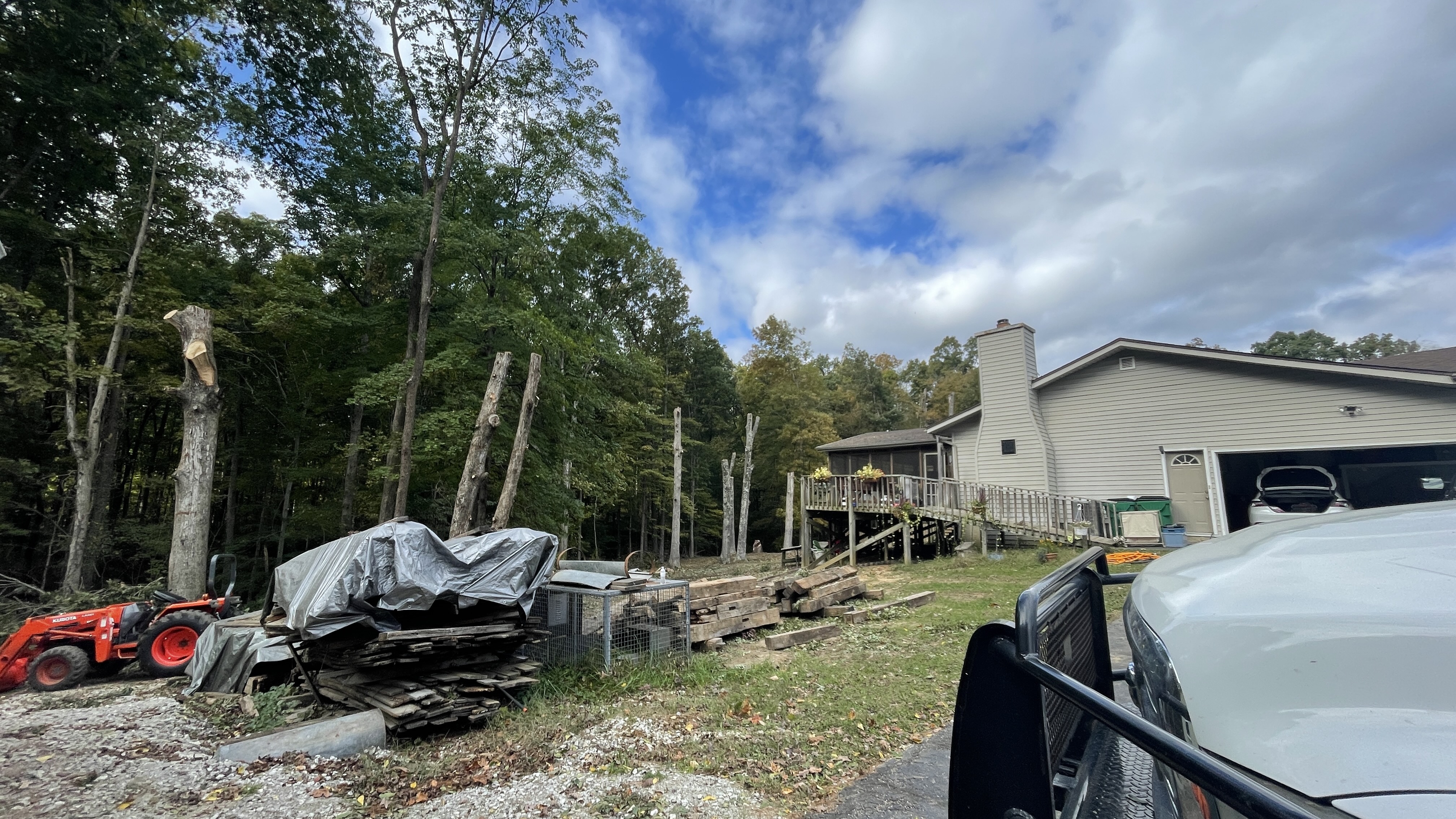 A residential property with a large house, a garage, and a wooded area. The yard appears to be undergoing some sort of construction or renovation, with a tractor, stacks of lumber, and tree stumps visible. The sky is partly cloudy.