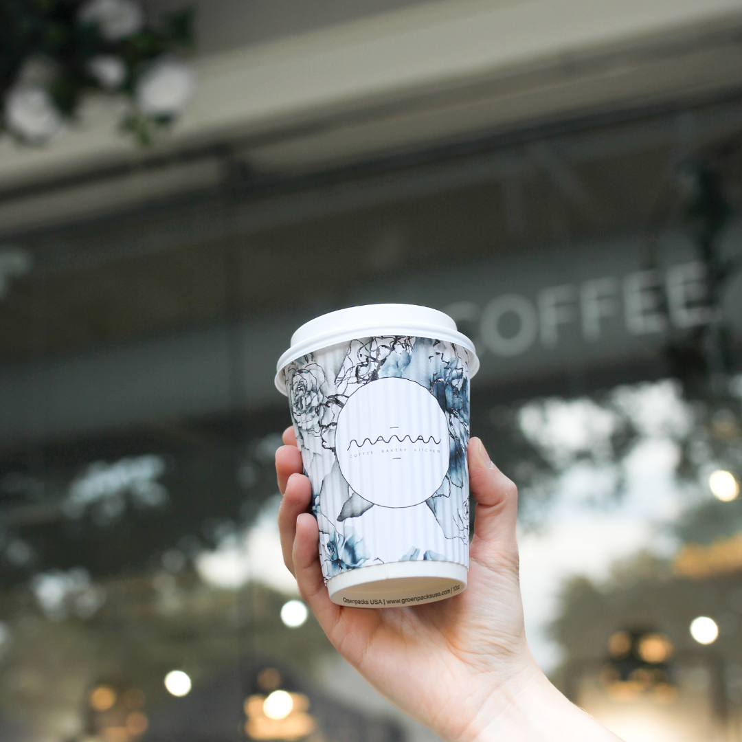 A branded maman takeaway coffee cup is held in front of the Park Cities café entrance. The floral cu 