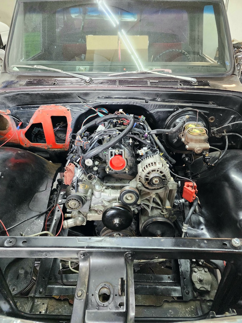 A close-up, eye-level shot captures the engine bay of a vehicle undergoing repair or modification. The focus is on the complex assembly of the engine, with various components, wiring harnesses, and hoses visible. The engine block is predominantly gray, with some black and metallic accents. A bright red cap is fitted to one of the engine's intake ports. To the right of the engine, a gold-colored master cylinder with attached brake lines is present. The surrounding area includes black fenders, red painted components on the left, and exposed metal framing at the bottom. The windshield is visible at the top, reflecting overhead fluorescent lights and showing two cardboard boxes within the vehicle's cabin.
