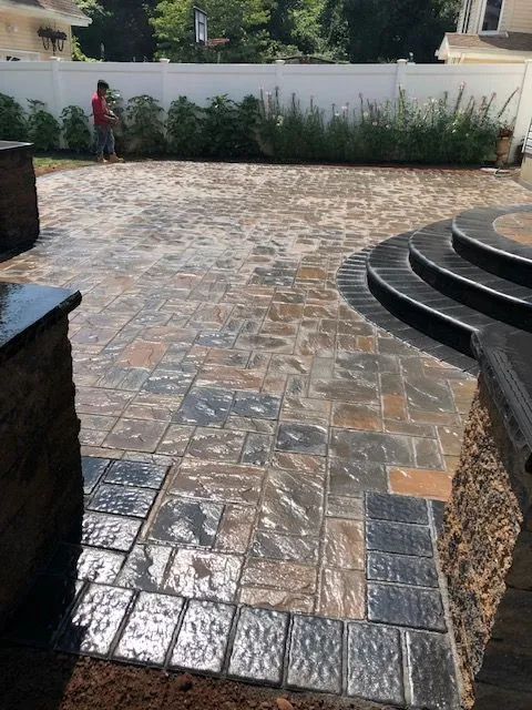 A person in a red shirt and blue jeans is watering a newly paved patio with stamped concrete. The patio has a curved staircase on the right side and is surrounded by a white vinyl fence and lush green plants. A basketball hoop is visible in the background.