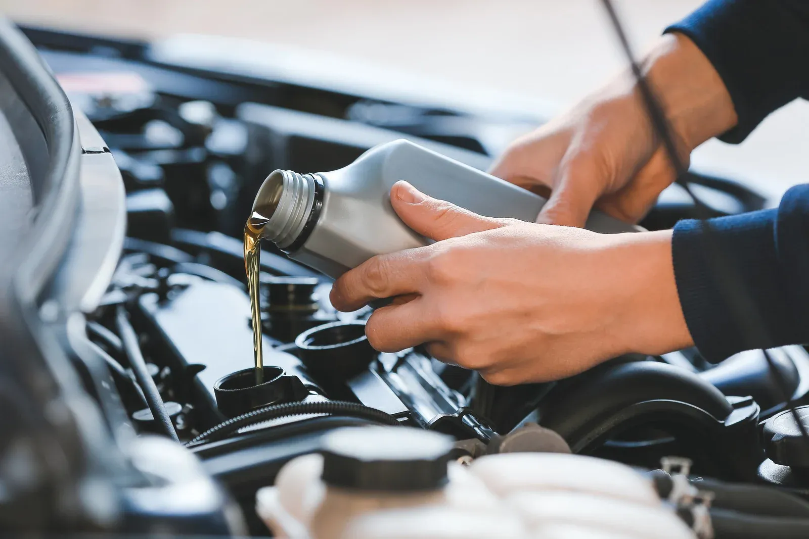 Hands pouring fresh engine oil into a car engine using a plastic container. The scene highlights routine vehicle maintenance, ensuring proper lubrication for engine components to improve performance, efficiency, and longevity in an automotive service setting.