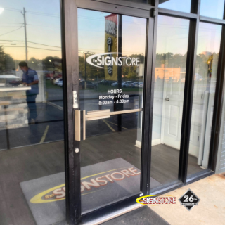 Entrance door of Signstore showing business hours: Monday - Friday, 8:00am - 4:30pm. The glass door has the Signstore logo, and the interior features a mat with the Signstore branding. The "26" anniversary badge is visible at the bottom.