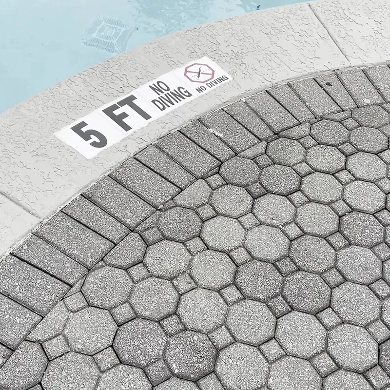A close-up view of a swimming pool edge showing a "5 FT NO DIVING" sign and the textured pavement surrounding the pool.