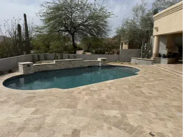 Custom travertine pool deck in Scottsdale with a stacked stone water feature wall and spillway. Wide decking gives the yard room to breathe, and the tumbled travertine finish stays cool underfoot even mid-summer. Built by Hacienda Outdoors, ROC #325952.