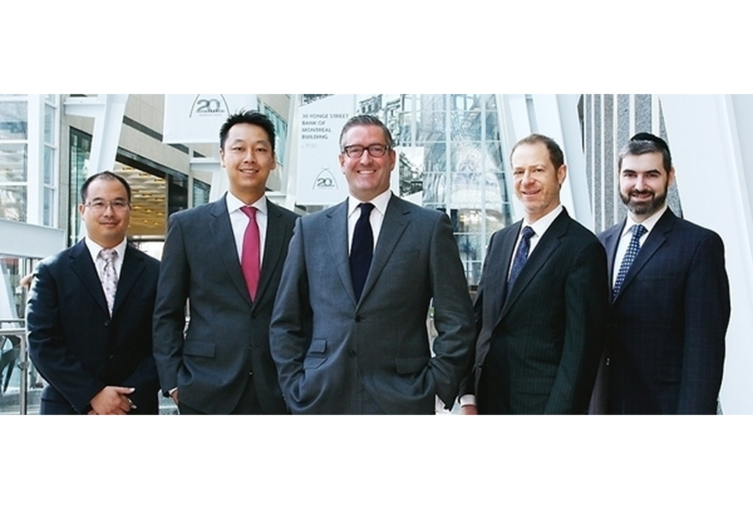 Five men in suits stand in a bright atrium, smiling confidently. Modern architecture and glass walls create a professional, welcoming atmosphere.