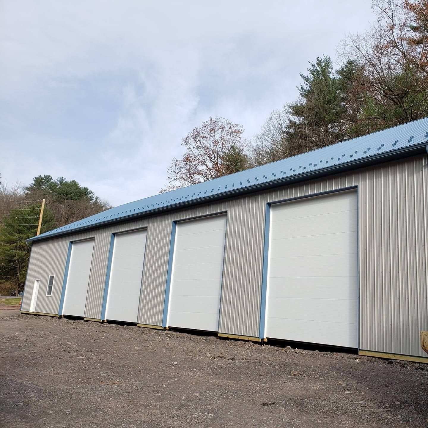 A long metal garage building with vertical siding, multiple large white roll-up doors, and blue trim, situated on a gravel surface with trees behind, under a cloudy sky.