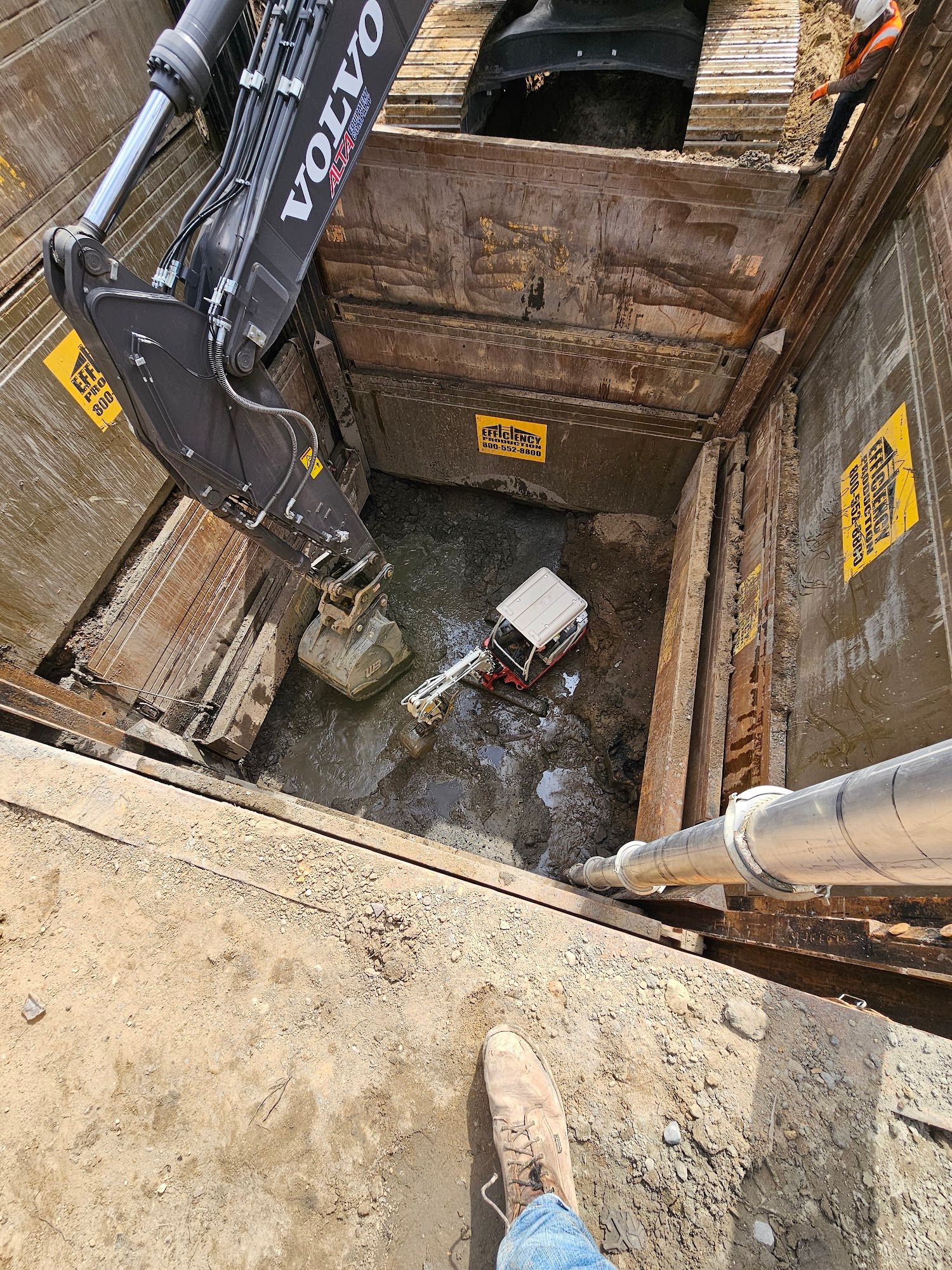 Excavation equipment operates within a deep shored trench while removing soil and groundwater. The project requires trench support systems to safely complete underground utility installation.