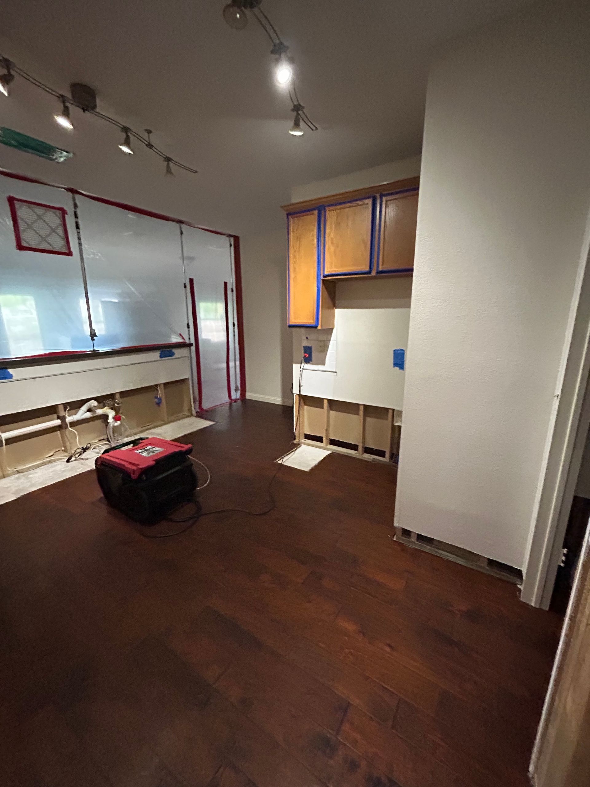 A partially restored kitchen shows dark hardwood flooring, exposed lower wall cavities, taped cabine 