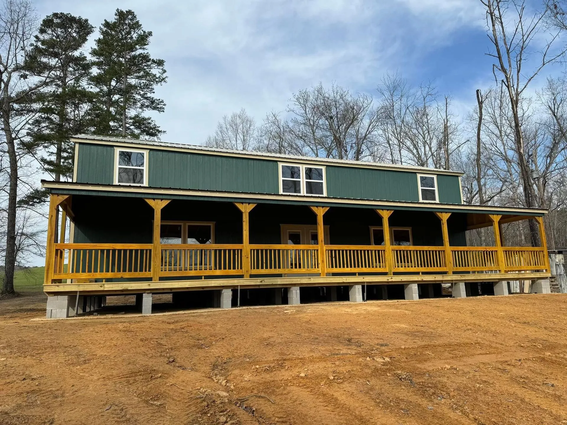 Completed elevated cabin featuring wraparound covered porch, green siding, and sturdy wood railings. Installed on concrete block foundation, providing spacious residential living in a peaceful outdoor environment.