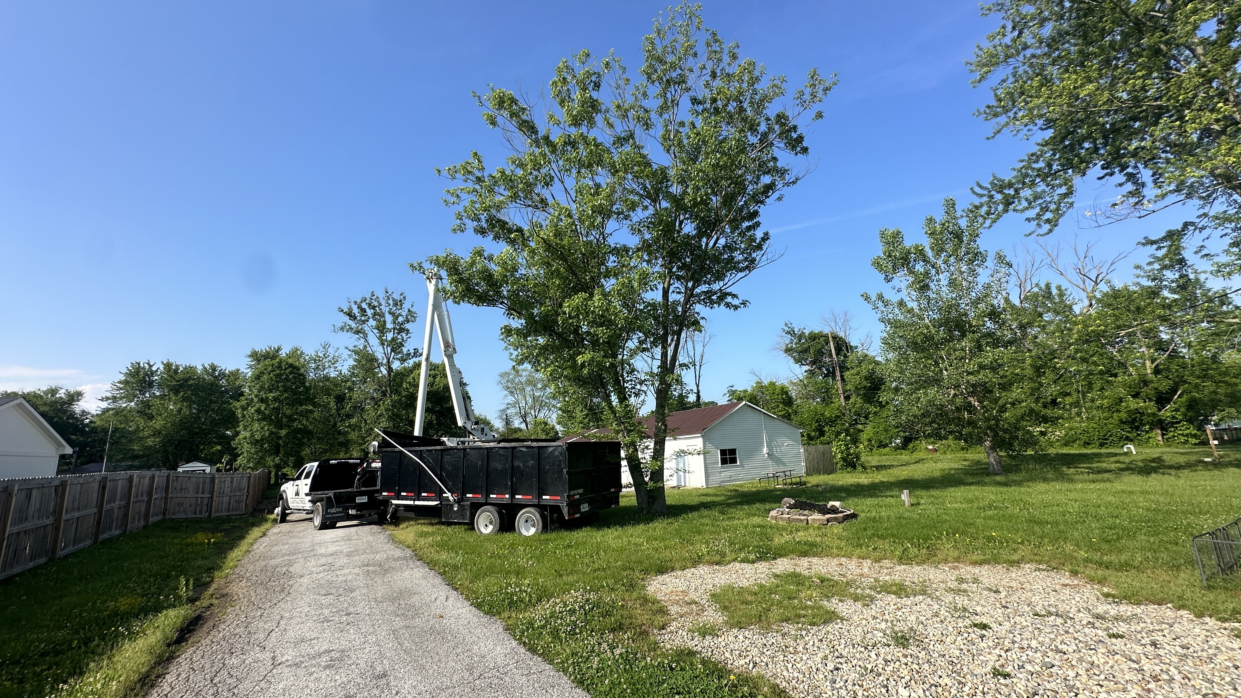 A white truck is parked next to a large black dumpster on a gravel driveway. A tall, white hydraulic lift is extended from the truck, reaching towards the trees in the background. A small white building with a brown roof and a window is visible behind the dumpster. The area is surrounded by lush green trees and a clear blue sky. A wooden fence runs along the side of the property.