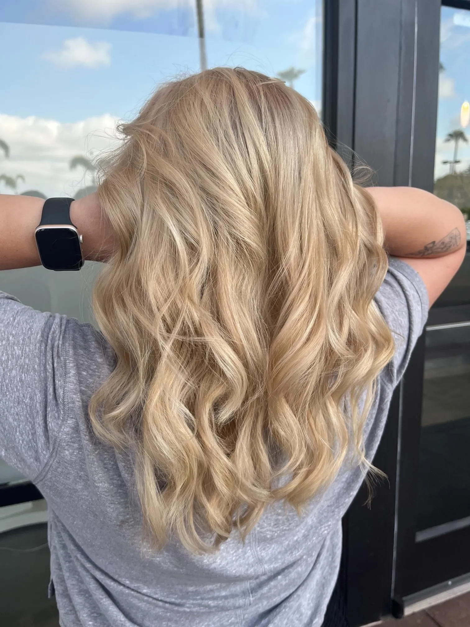 Medium-length blonde hair styled in soft beach waves with natural highlights and smooth texture. The hairstyle is photographed from the back, showcasing professional salon coloring and styling with bright dimension.
