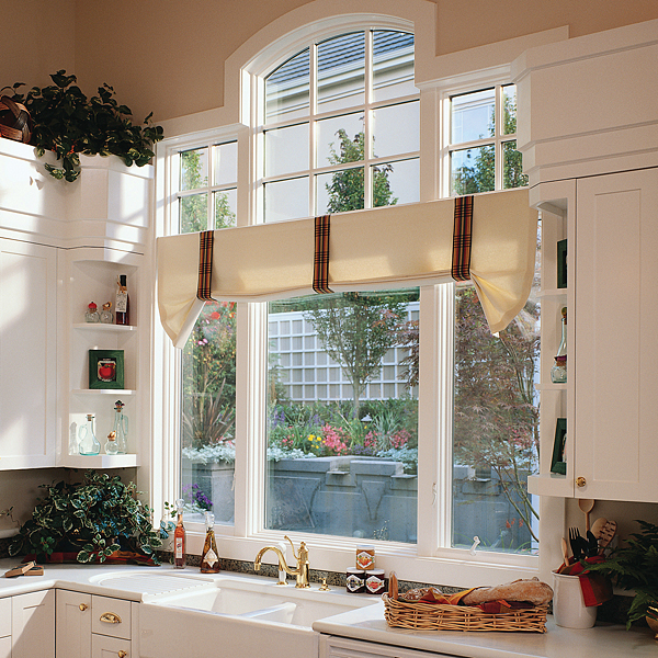 Bright kitchen featuring a large, white-framed window with an arched upper section above a sink. The window overlooks greenery and a landscaped patio area. White cabinetry, a farmhouse-style sink, gold-toned hardware, plants, and decorative items surround the window.