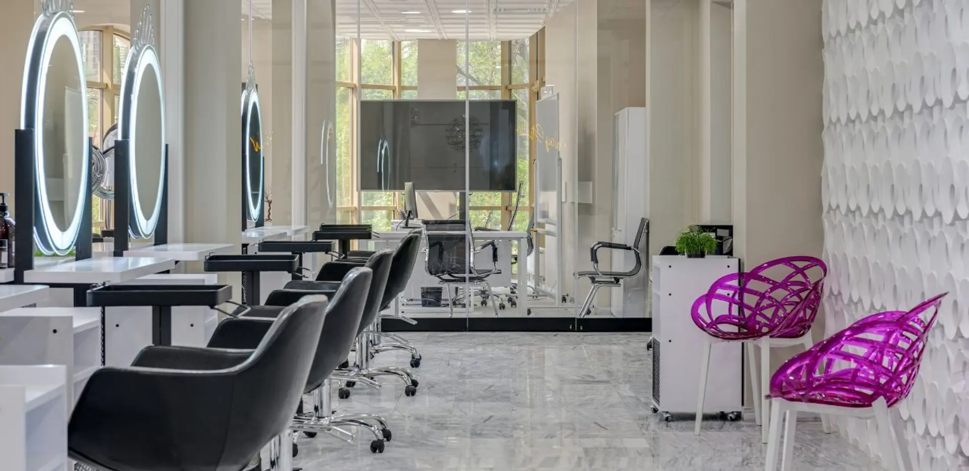 A modern, well-lit beauty salon with rows of styling stations, each equipped with a lighted mirror and chair. The salon features a minimalist design with white countertops and sleek black chairs. In the background, a large screen is mounted on the wall, and large windows offer a view of greenery. Two eye-catching purple chairs with intricate woven designs are placed near a textured white wall, adding a pop of color to the space.