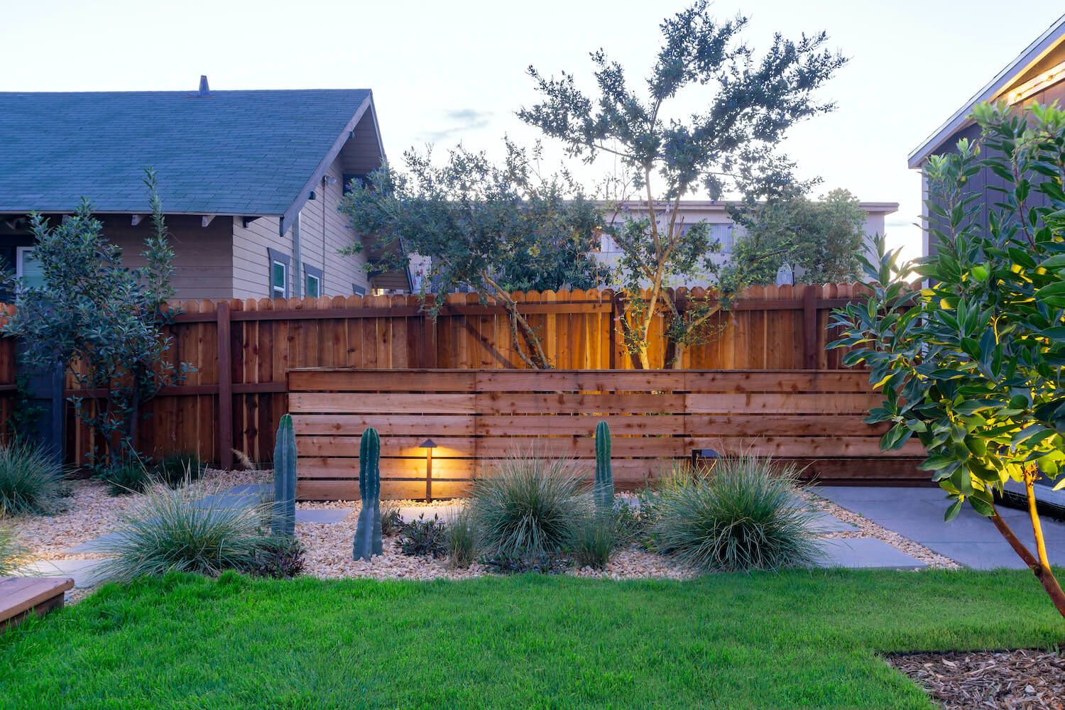 Landscaped backyard with lush lawn, cacti, ornamental grasses, warm garden lighting, trees, and a wooden privacy fence at dusk.