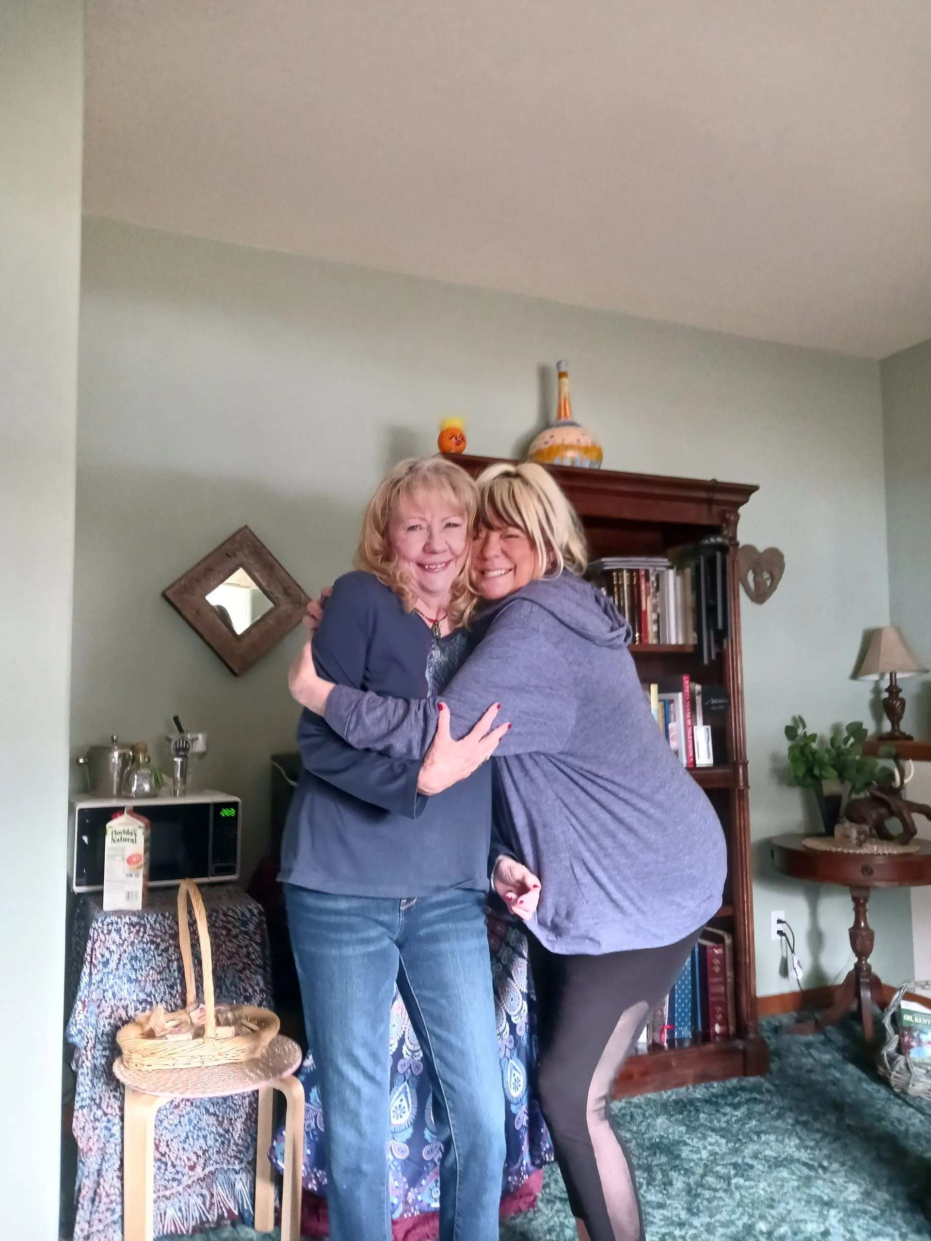 Two smiling women share a warm embrace inside a cozy living room with bookshelves, decorative furniture, and soft lighting. Casual clothing and cheerful expressions create a relaxed atmosphere while home décor, baskets, and plants add comfort and personal character.