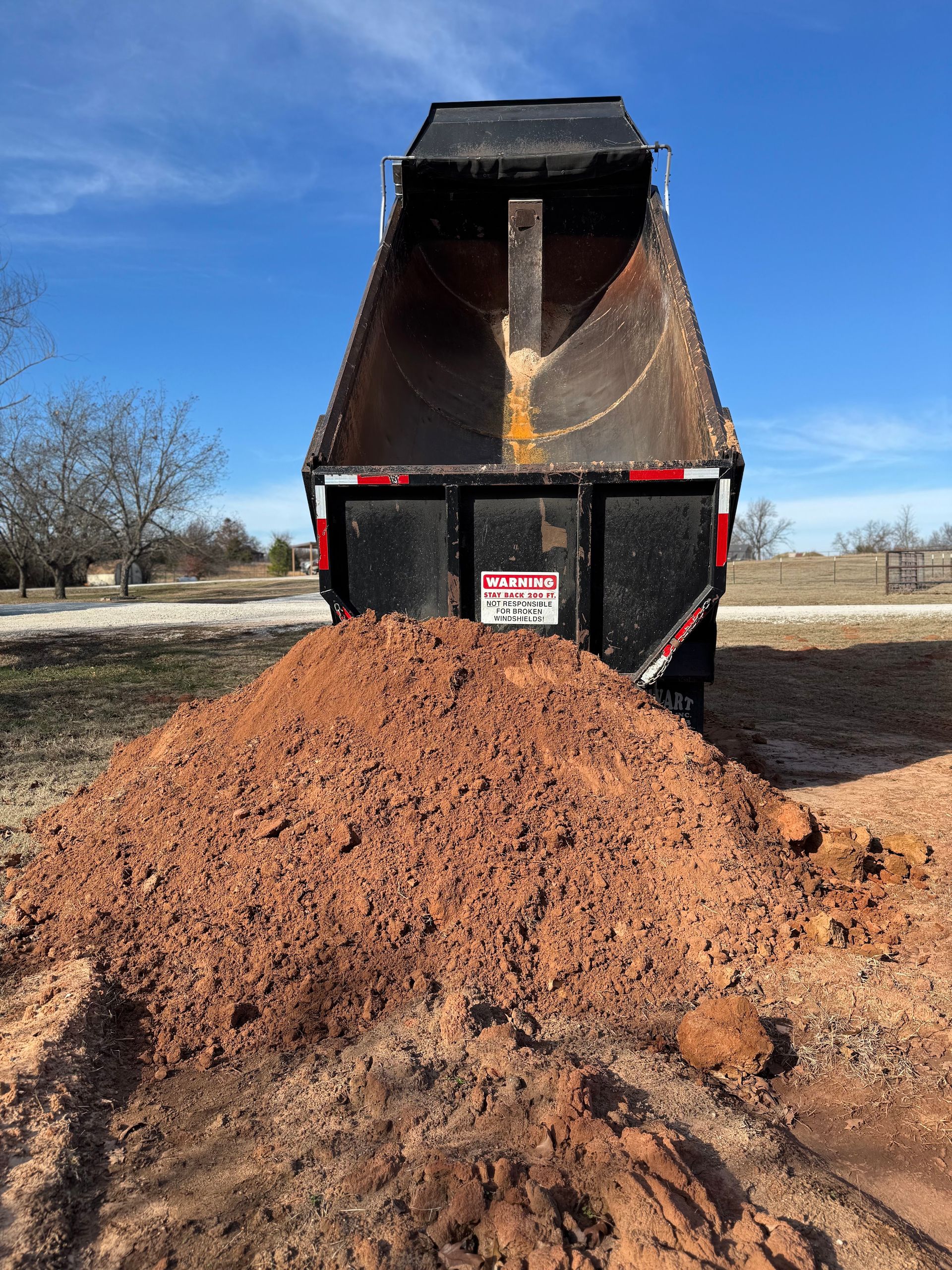 L & R Logistics LLC dump truck unloading a large pile of reddish soil outdoors, demonstrating material hauling and delivery capabilities for construction, landscaping, and property improvement projects across Oklahoma.