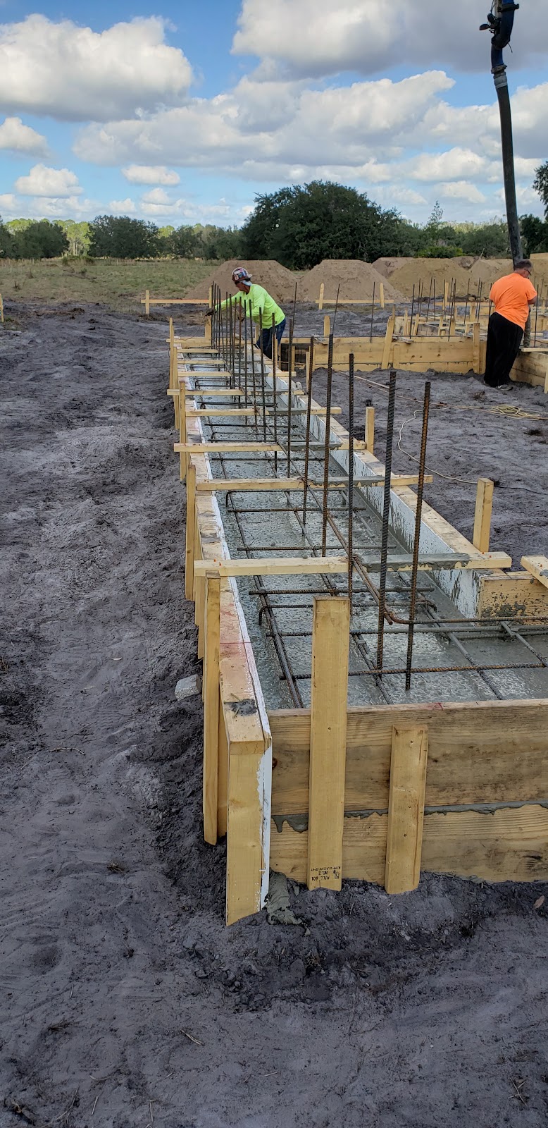 Construction workers are pouring concrete into a foundation form. A concrete pump hose is visible ov 