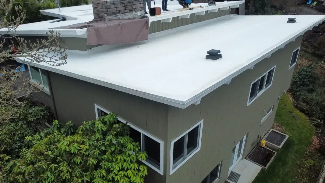 An aerial shot of a two-story, modern-style home with a flat roof. Several workers are visible on the white roof, possibly performing a new installation or repair job.