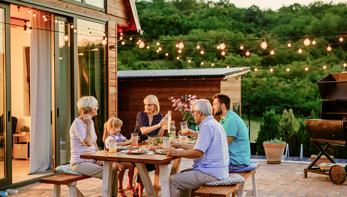 A multigenerational family having dinner on a patio outside a cottage.