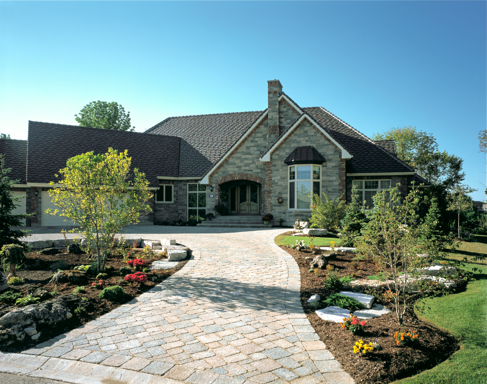 Stone country house with peaked roofs, arched entry, landscaped gardens, flowering plants, and a curved brick walkway under blue skies.