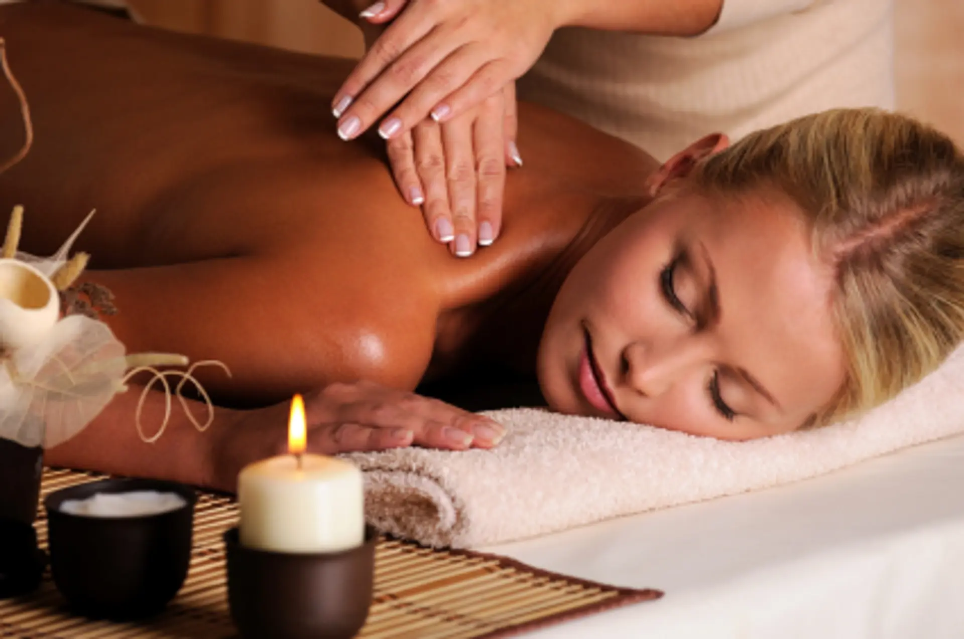 A close-up shot shows a woman receiving a back massage with her eyes closed. Her body is oiled, and her skin has a warm, glowing tone. A masseuse's hands are visible massaging her shoulders. In the foreground, there is a lit candle in a dark holder and a small black bowl filled with what appears to be lotion or oil, placed on a bamboo mat. The setting suggests a tranquil spa or wellness environment.