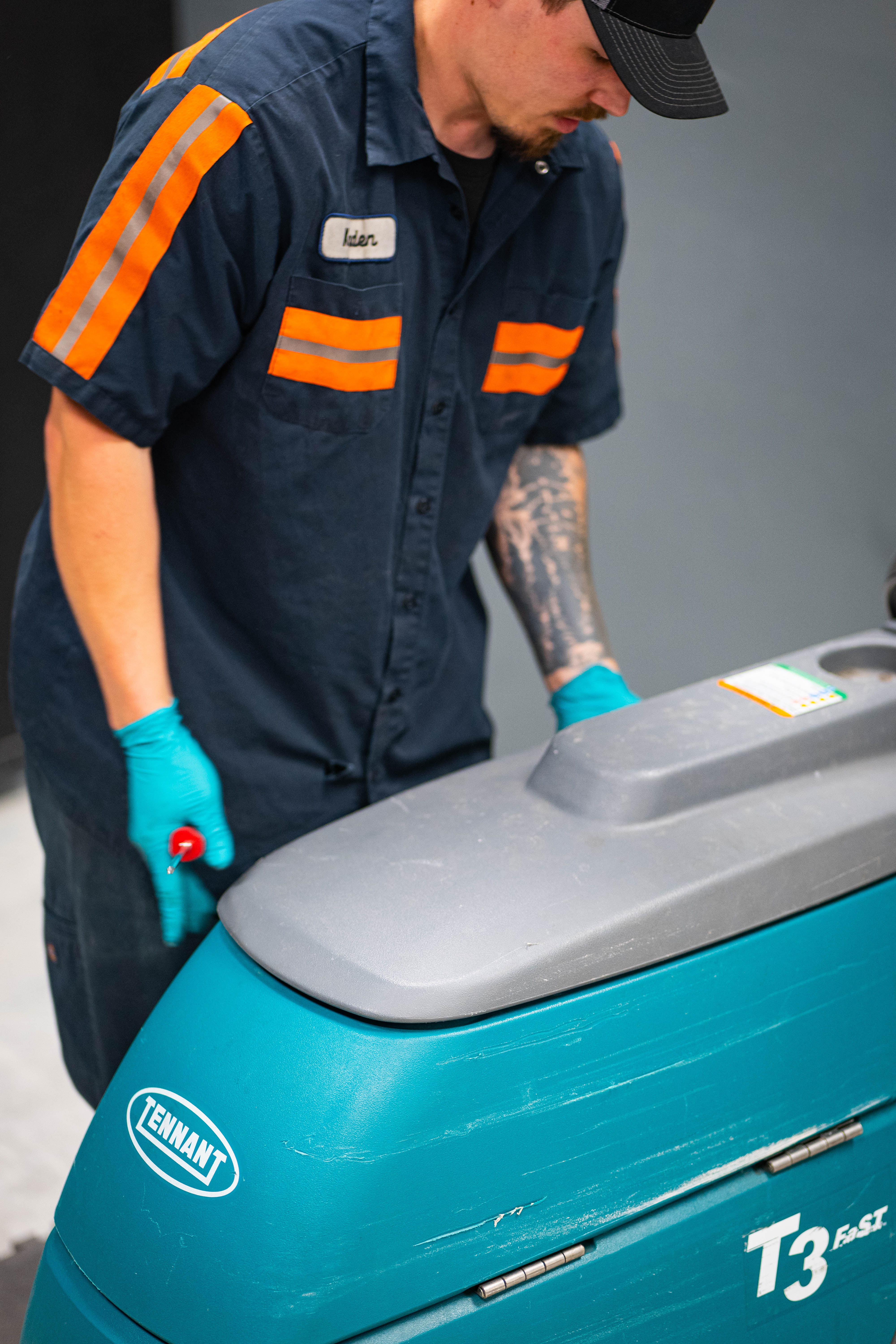 A certified technician from WAY Solutions performing a visual inspection and routine maintenance on the upper tank and control area of a teal Tennant T3 walk-behind floor scrubber.