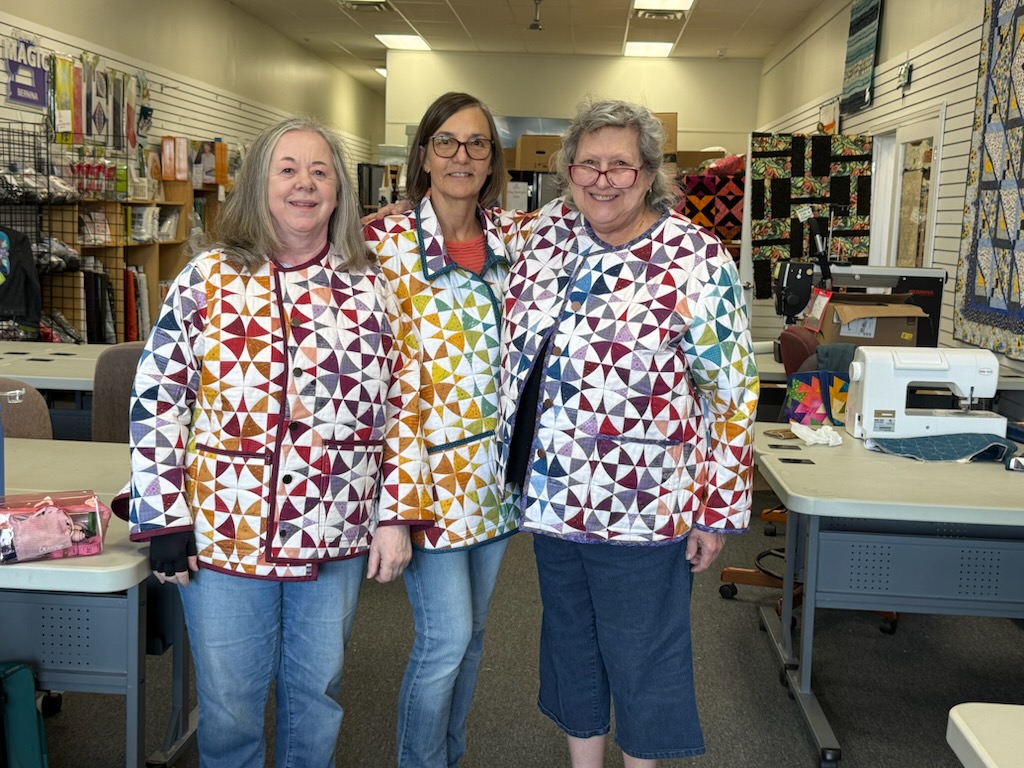 Three quilters proudly wear their handmade quilted jackets during a Decorative Stitch event. Their colorful creations showcase expert piecing, decorative stitching, and the creativity inspired through quilting classes.