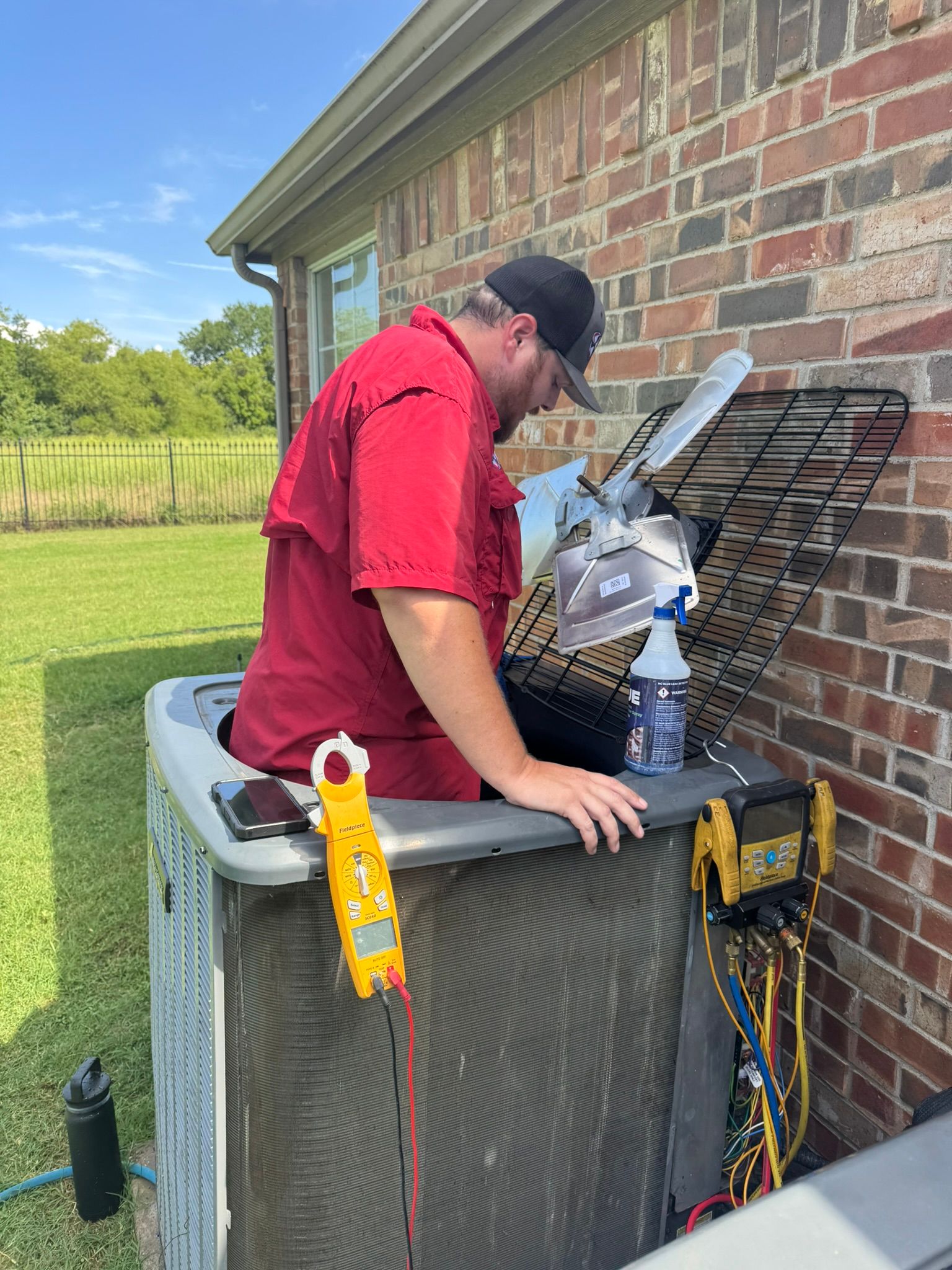 An HVAC technician in a red shirt is leaning into a large outdoor air conditioner unit, performing a maintenance check. Tools, including a clamp meter, are visible on the unit.