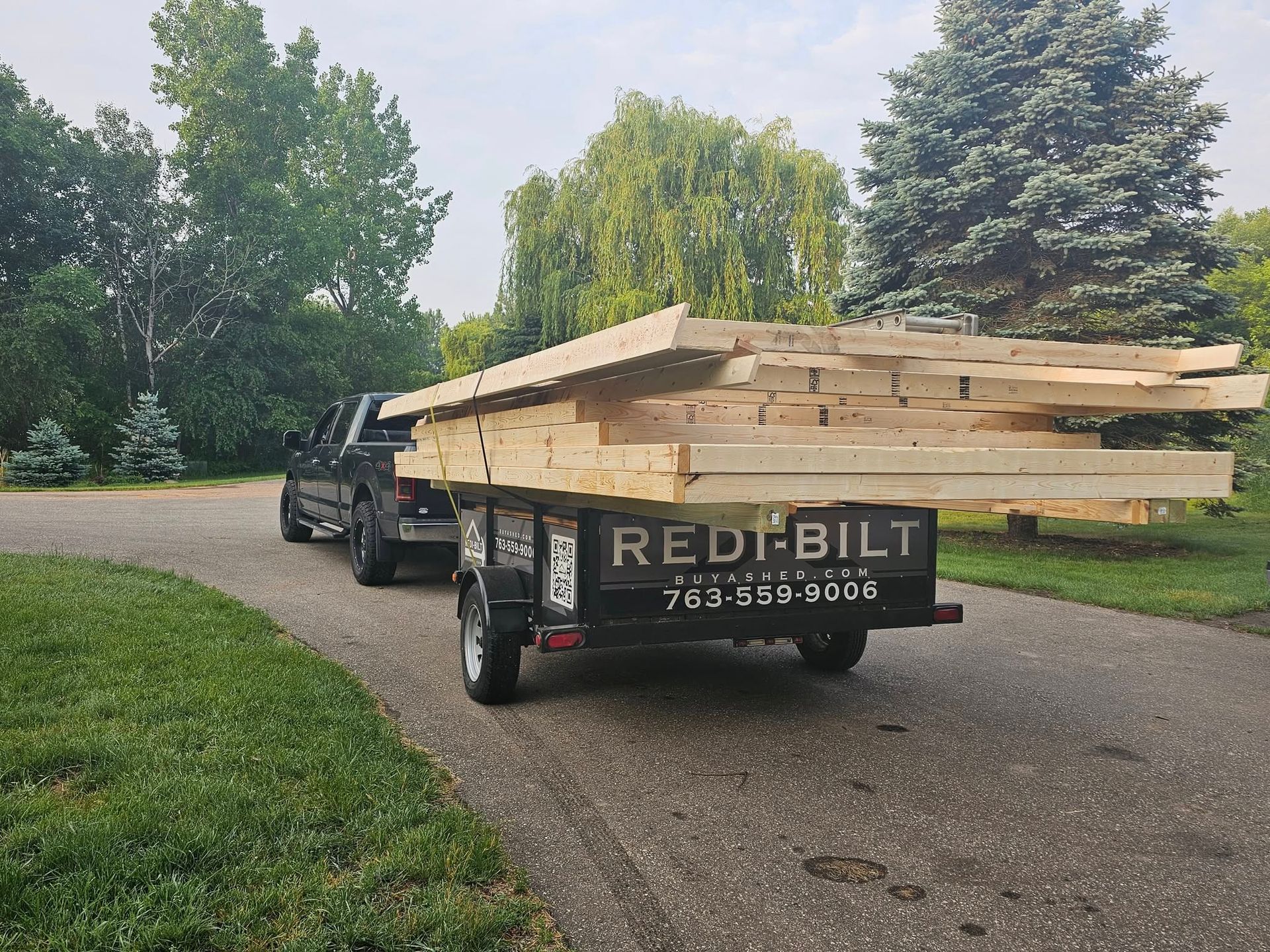 A black pickup truck is towing a dark utility trailer loaded with a large stack of dimensional lumber, likely for a shed build. The trailer features the "REDI-BILT" logo, website, and phone number on the back panel.